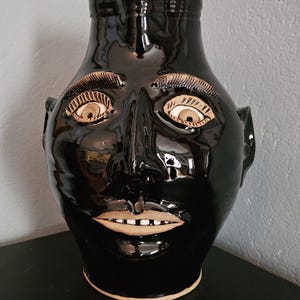 May include: A glossy black ceramic face jug featuring a sculpted human face with prominent eyebrows, wide eyes, and a mouth with visible teeth. The jug has a rounded body, a narrow neck, and a small spout at the top, with unglazed tan clay accents.