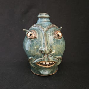 May include: A ceramic bottle with a face design. The bottle is glazed in shades of blue and green, with a detailed face featuring eyes, a nose, and a smiling mouth. The bottle has a small neck and two small ear-like protrusions on the sides.