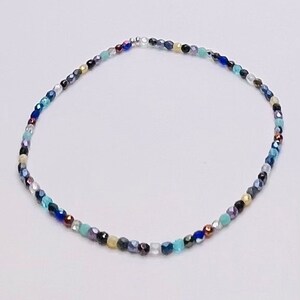 May include: A delicate bracelet made of small, colorful beads. The beads are in shades of blue, green, gold, and silver. The bracelet is strung on a clear elastic cord.