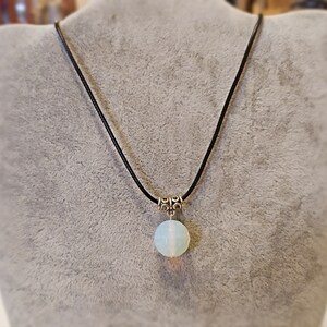 May include: A white opal pendant necklace on a black cord. The pendant is round and has a silver clasp.