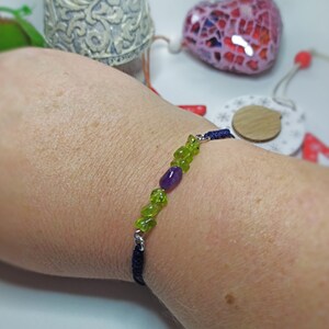 May include: A dark blue cord bracelet with green and purple gemstone beads. The bracelet is tied with a knot and has a silver clasp.
