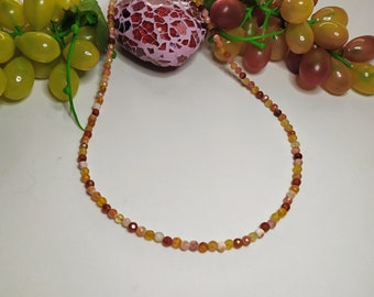 May include: A necklace made of small, round, multi-colored beads. The beads are shades of red, orange, and yellow. The necklace is lying on a white surface.