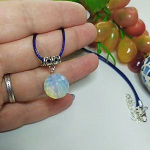 May include: A white opal gemstone pendant necklace with a silver chain and a blue cord. The pendant is round and has a faceted surface.