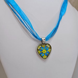 May include: A blue ribbon necklace with a heart-shaped pendant featuring a yellow and blue floral design.