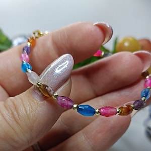 Puede incluir: Un brazalete colorido con cuentas doradas y cuentas de piedras preciosas de varios colores, incluyendo rosa, azul, marrón y morado.