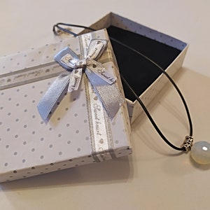 May include: A white gift box with a silver ribbon and polka dots. The box is open and a black cord necklace with a white opal pendant is inside.