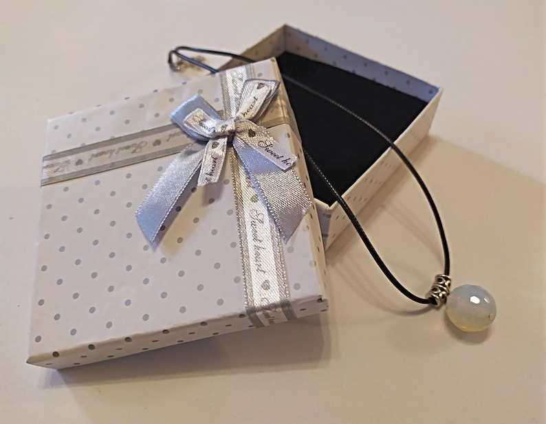 May include: A white gift box with a silver ribbon and polka dots. The box is open and a black cord necklace with a white opal pendant is inside.