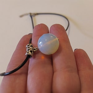 May include: A white opal gemstone pendant on a black cord necklace. The pendant is round and has a silver clasp.