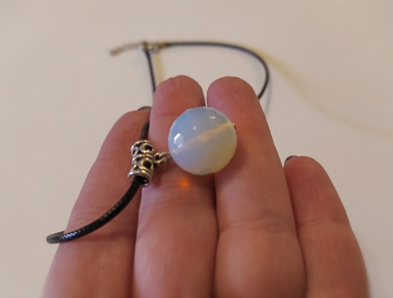 May include: A white opal gemstone pendant on a black cord necklace. The pendant is round and has a silver clasp.