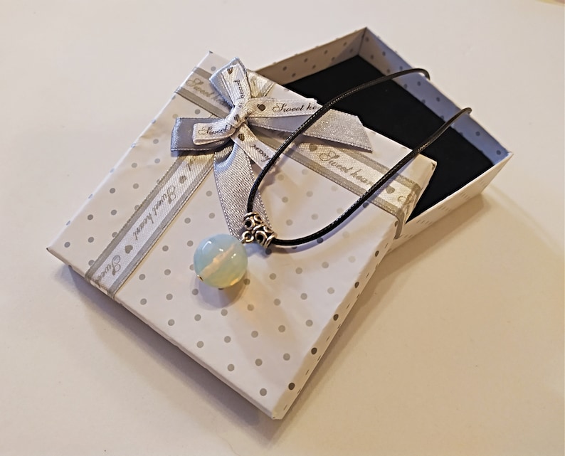 May include: A white gift box with a silver ribbon and a white opal pendant necklace with a black cord. The box has a white background with black polka dots and the text "Sweet heart" on the ribbon.