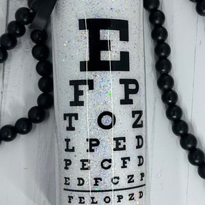 May include: A white glitter tumbler with a black eye chart design. The eye chart is printed in black and includes the letters E, F, P, T, O, Z, L, P, E, D, and C. A black ribbon is attached to the top.