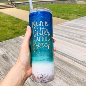 May include: A blue and green glitter tumbler with a white base and gold glitter accents. The tumbler has the text "Life is better at the beach" with a starfish design.