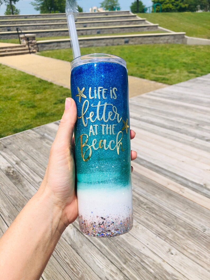 May include: A blue and green glitter tumbler with a white base and gold glitter accents. The tumbler has the text "Life is better at the beach" with a starfish design.