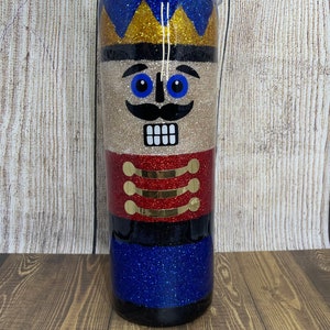 May include: A glitter tumbler with a nutcracker design. The tumbler is decorated with gold, red, blue, and black glitter. The nutcracker has a gold crown, blue eyes, a black mustache, and a red and gold uniform.