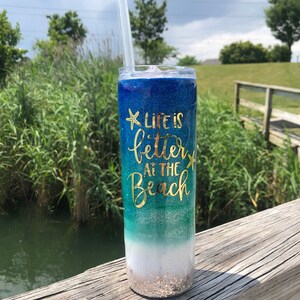 May include: A blue, green, and white glitter tumbler with a gold glitter bottom. The tumbler has a straw and says "Life is better at the beach" with a gold glitter starfish on each side of the text.