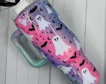 May include: A glittery tumbler with a handle, featuring a Halloween theme. The tumbler has a gradient of pink, purple, and blue with white ghost and black bat designs. The handle is a teal color. A black ribbon is attached to the lid.