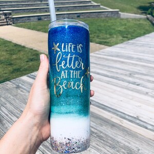 May include: A blue and green glitter tumbler with a white base. The tumbler has a gold glitter design and the text "Life is better at the beach" with a gold glitter starfish on either side of the text.