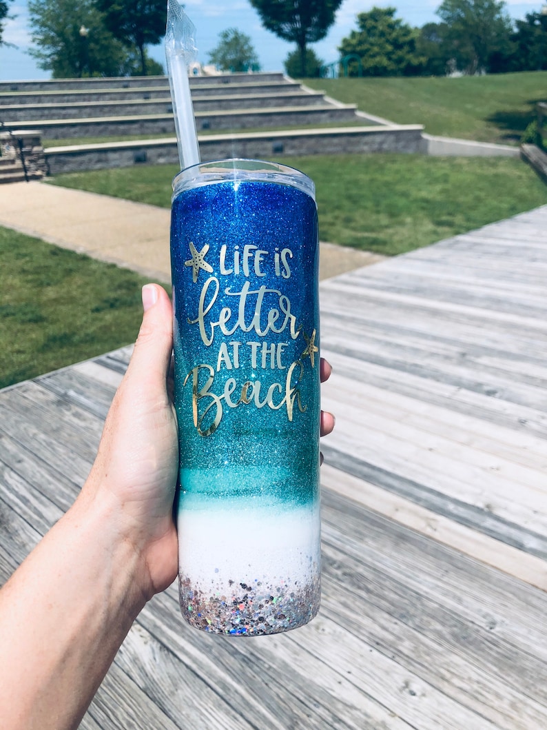 May include: A blue and green glitter tumbler with a white base. The tumbler has a gold glitter design and the text "Life is better at the beach" with a gold glitter starfish on either side of the text.