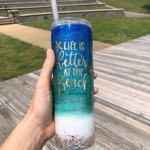 May include: A blue, green, and white glitter tumbler with a straw. The tumbler has the text "Life is better at the beach" in gold lettering.