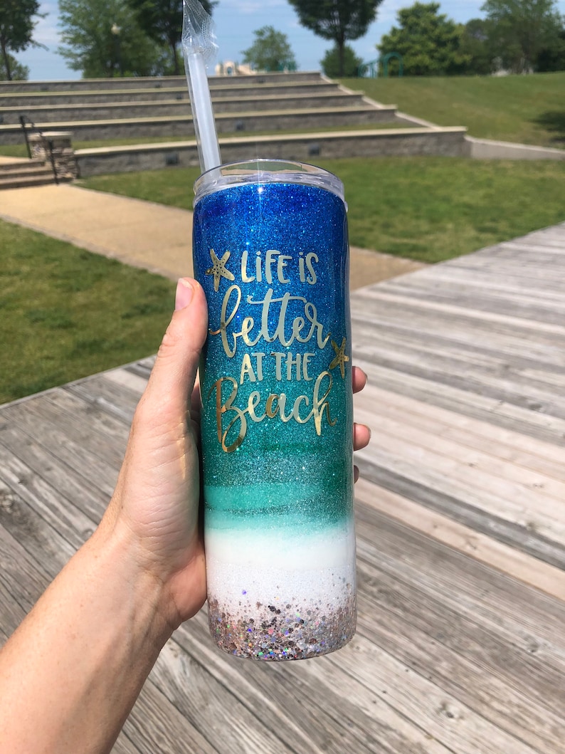 May include: A blue, green, and white glitter tumbler with a straw. The tumbler has the text "Life is better at the beach" in gold lettering.