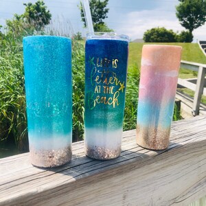 May include: Three glitter tumblers with a beach theme. The tumbler on the left is blue and green with a white sandy beach at the bottom. The middle tumbler is blue and green with the text "Life is better at the beach" and a gold starfish. The tumbler on the right is pink, blue, and gold with a white sandy beach at the bottom.