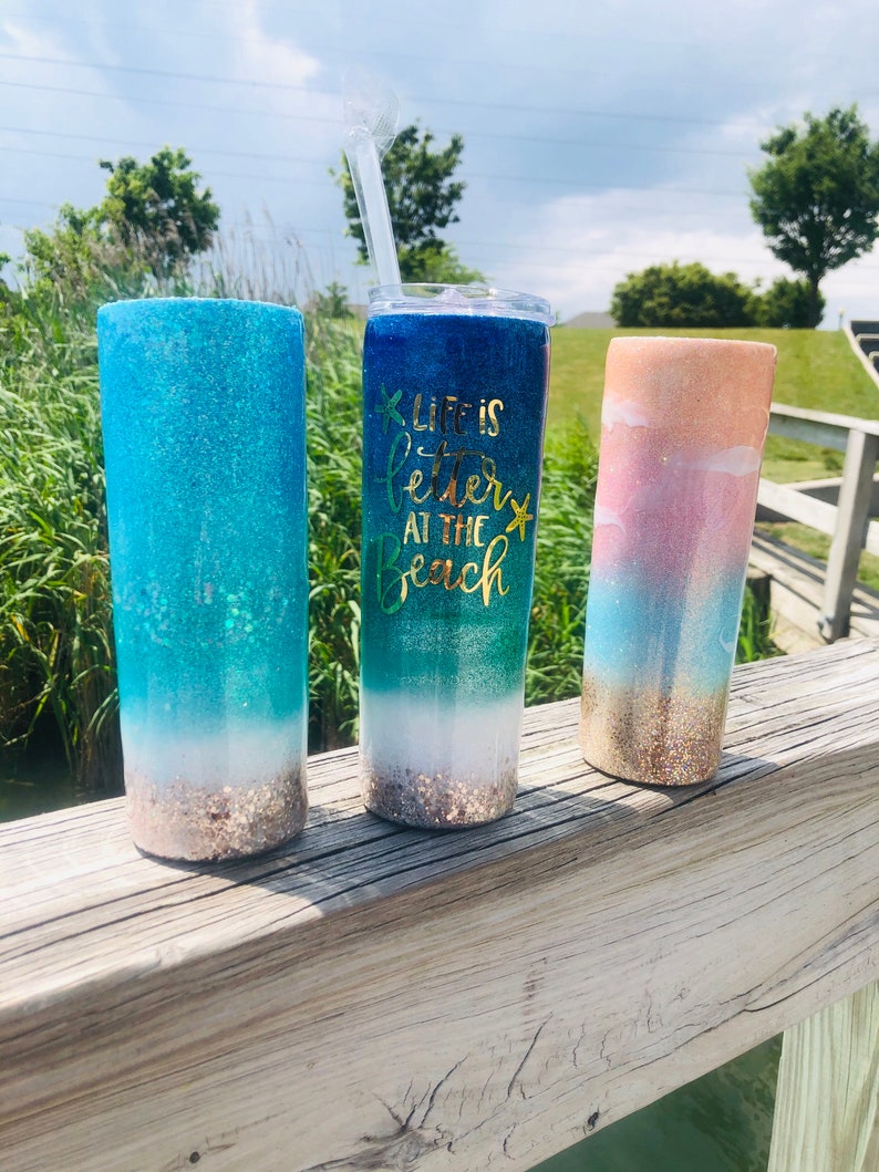 May include: Three glitter tumblers with a beach theme. The tumbler on the left is blue and green with a white sandy beach at the bottom. The middle tumbler is blue and green with the text "Life is better at the beach" and a gold starfish. The tumbler on the right is pink, blue, and gold with a white sandy beach at the bottom.