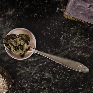 May include: A close-up of a green marble surface with a small glass jar filled with dried green herbs, a small white dish with a silver spoon containing more dried herbs, and a small gold-colored rock.