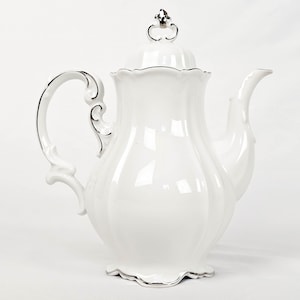 May include: A white porcelain teapot with a silver trim and handle. The teapot has a curved spout and a decorative lid with a silver finial. The handle has an ornate design. The base of the teapot has a scalloped edge.