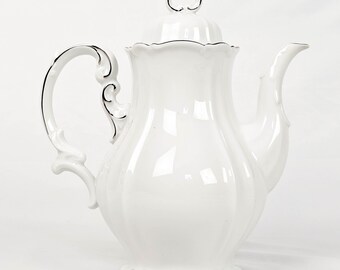 May include: A white porcelain teapot with a silver trim and handle. The teapot has a curved spout and a decorative lid with a silver finial. The handle has an ornate design. The base of the teapot has a scalloped edge.