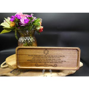 May include: A rectangular wooden tray with engraved Hebrew text and decorative elements. The tray is made of light brown wood and is displayed on a wooden surface. A vase of flowers is in the background.