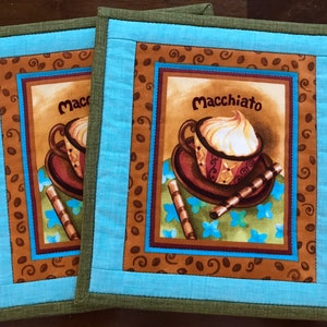 May include: Two brown and blue quilted coffee cup coasters with a brown and white coffee cup with whipped cream and the text "Macchiato" on the coaster.