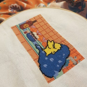 May include: Cross-stitch embroidery of a yellow and blue cartoon character in a blue and white tiled bathroom setting. The character is in a bathtub with a bottle of shampoo and a bar of soap.
