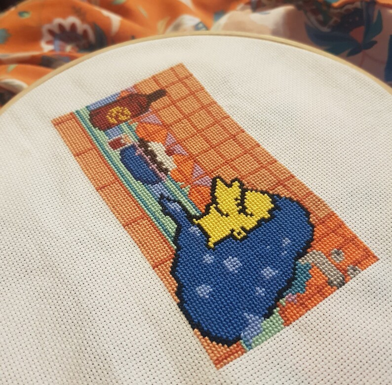 May include: Cross-stitch embroidery of a yellow and blue cartoon character in a blue and white tiled bathroom setting. The character is in a bathtub with a bottle of shampoo and a bar of soap.