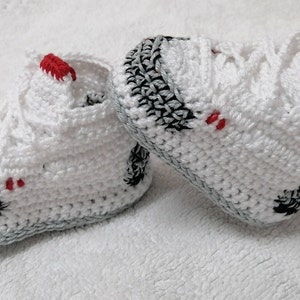 May include: A pair of white crocheted baby shoes with black and gray accents and red trim.