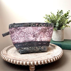 May include: A small, patterned cosmetic bag with a zipper closure. The bag features a floral design in shades of purple and gray, with a black lace trim. It sits on a decorative wooden tray.