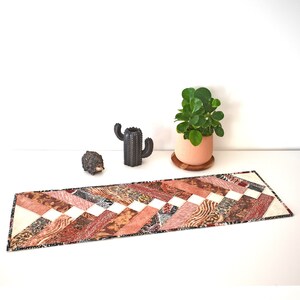 May include: A handmade table runner with a patchwork design. The runner features a repeating pattern of diagonal stripes in shades of brown, cream, and coral. The edges are finished with a black border. A decorative cactus and a potted plant are in the background.