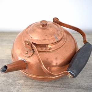 May include: A vintage copper tea kettle with a wooden handle. The kettle has a shiny, aged copper finish and a rounded body.