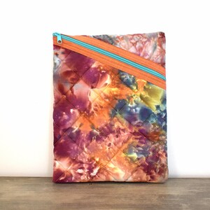 May include: Quilted fabric case with a vibrant tie-dye pattern in shades of orange, purple, and yellow. The case features a diagonal orange zipper with a turquoise zipper pull. The fabric has a diamond-shaped quilted design.