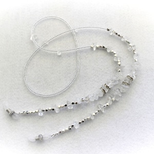 Puede incluir: Una delicada cadena para gafas con cuentas claras de forma irregular y pequeñas cuentas plateadas ensartadas en un fino cordón plateado. La cadena tiene bucles en cada extremo para sujetar las gafas. Diseño sencillo y elegante.