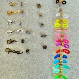 Puede incluir: Surtido de porta-cadenas para gafas de colores con detalles en metal dorado. Las cadenas son de plástico transparente y vienen en una variedad de colores, incluyendo negro, blanco, amarillo, rosa, rojo, azul, verde y turquesa. Las cadenas están unidas a cierres de metal dorado.
