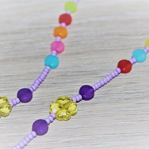 Puede incluir: Un collar de cuentas de colores con cuentas moradas y abalorios de flores amarillas. El collar presenta un arcoíris de colores, incluyendo rojo, naranja, amarillo, verde, azul y morado.