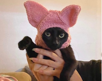 May include: A black cat wearing a pink knitted hat with large ears. The cat is being held by a person's hand.