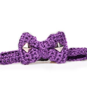 May include: A purple crocheted bow tie with silver pyramid studs. The bow tie is made of a thick, textured yarn and has a matching adjustable strap. The bow tie is set against a white background.