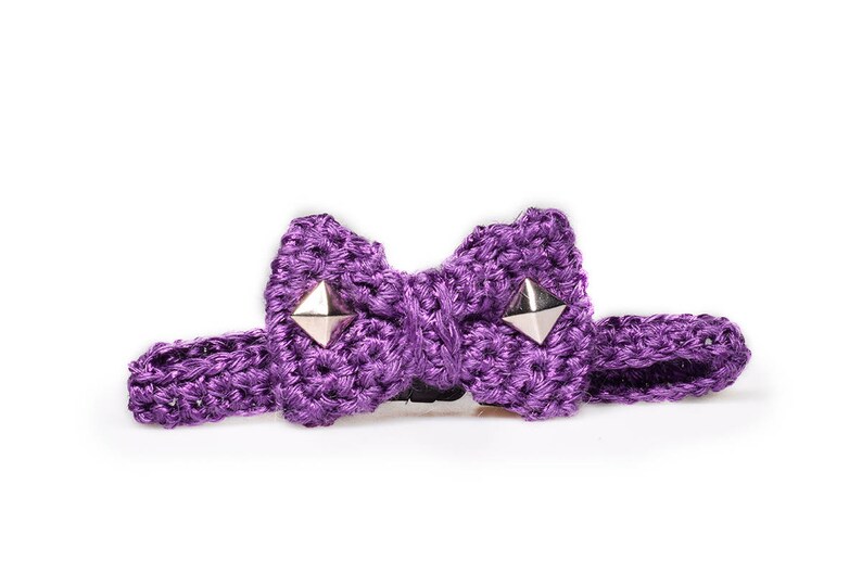 May include: A purple crocheted bow tie with silver pyramid studs. The bow tie is made of a thick, textured yarn and has a matching adjustable strap. The bow tie is set against a white background.