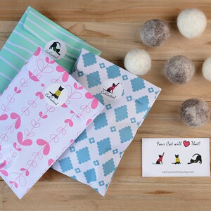 May include: A wooden surface displays cat toys and packaging. Three patterned envelopes with stickers are visible. Six felt balls in grey and white are also present. A small card with cat illustrations is included in the image.