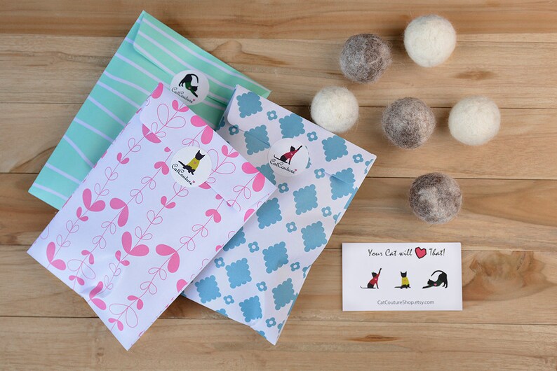 May include: A wooden surface displays cat toys and packaging. Three patterned envelopes with stickers are visible. Six felt balls in grey and white are also present. A small card with cat illustrations is included in the image.