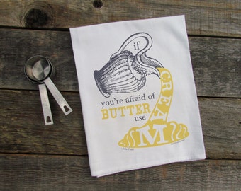May include: A white kitchen towel with a yellow and black illustration of a pitcher pouring butter. The text reads "If you're afraid of butter, use more."  The quote is attributed to Julia Child.