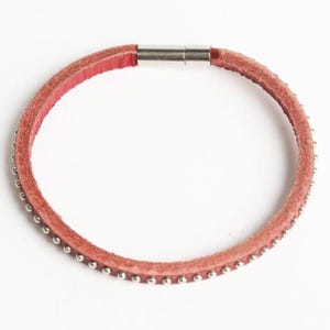 May include: A thin, circular bracelet made of reddish-brown leather with a textured surface. The outer edge of the leather band is lined with small, evenly spaced silver-colored metal studs. It features a cylindrical silver-tone metal magnetic clasp.