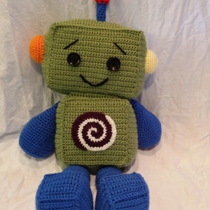 Pode incluir: Um brinquedo de robô de crochê verde com braços e pernas azuis. O robô tem um rosto sorridente com olhos pretos e uma boca preta. O robô tem uma antena vermelha, azul e laranja no topo da cabeça. O robô tem um design em espiral roxo e branco no peito.