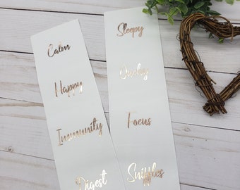 Puede incluir: Dos tarjetas blancas con letras doradas rosadas. Las palabras en las tarjetas son "Calm", "Happy", "Immunity", "Digest", "Sleepy", "Queasy", "Focus" y "Shiffles".
